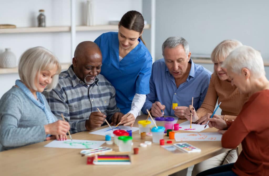 The Legacy At Falcon Point |  A group of seniors sitting on a table holding a small flat brush and painting, while being supervised by a nurse