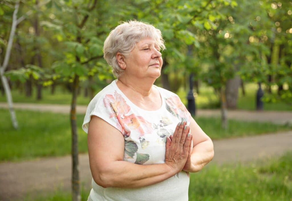 The Legacy At Falcon Point | Senior woman in nature practising gentle breathing and meditation with her hands together in front of her