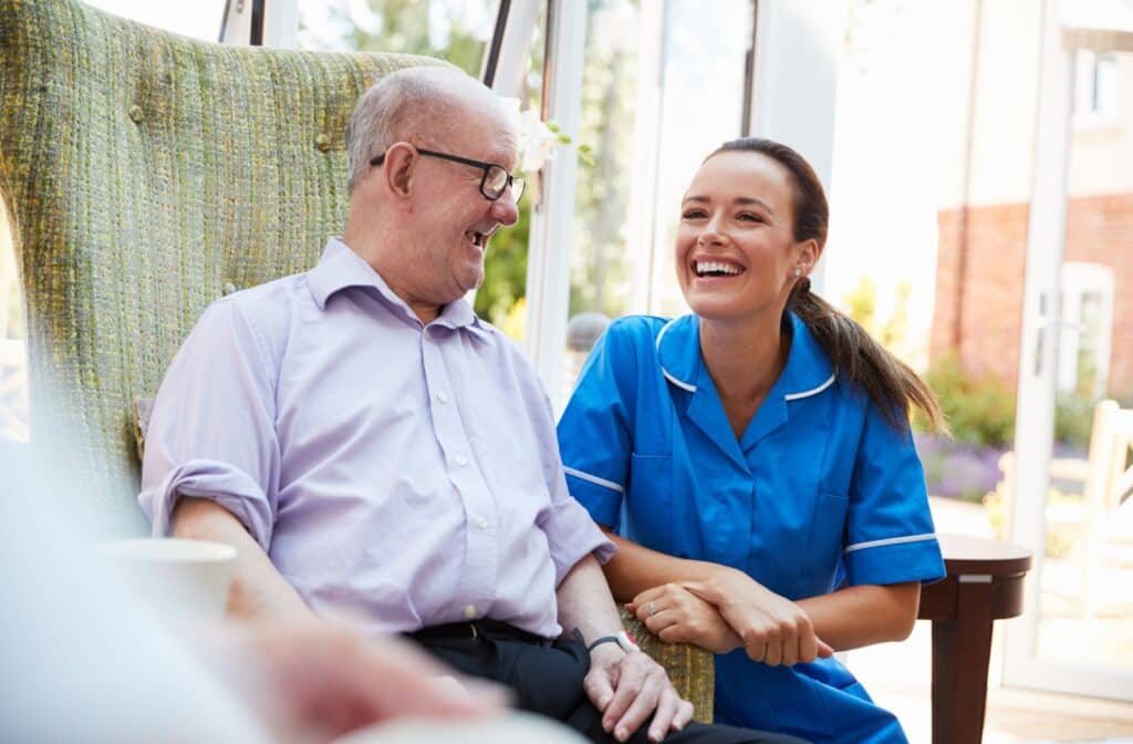 The Legacy At South Plains | An older adult man in a memory care facility sitting on a chair smiling and having a conversation with a nurse.