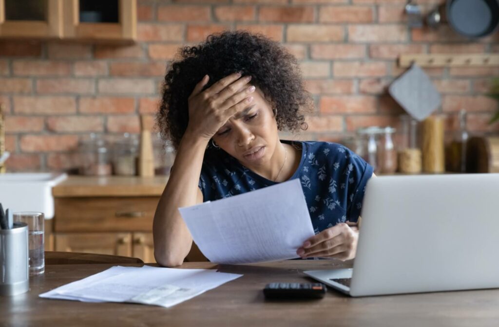 A stressed person holds their head as they review a financial document next to their laptop.