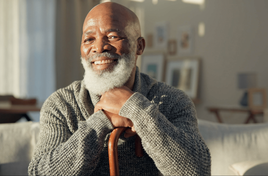 The Legacy at South Plains | A senior rests both hands on a wooden cane, smiling bright in their home.