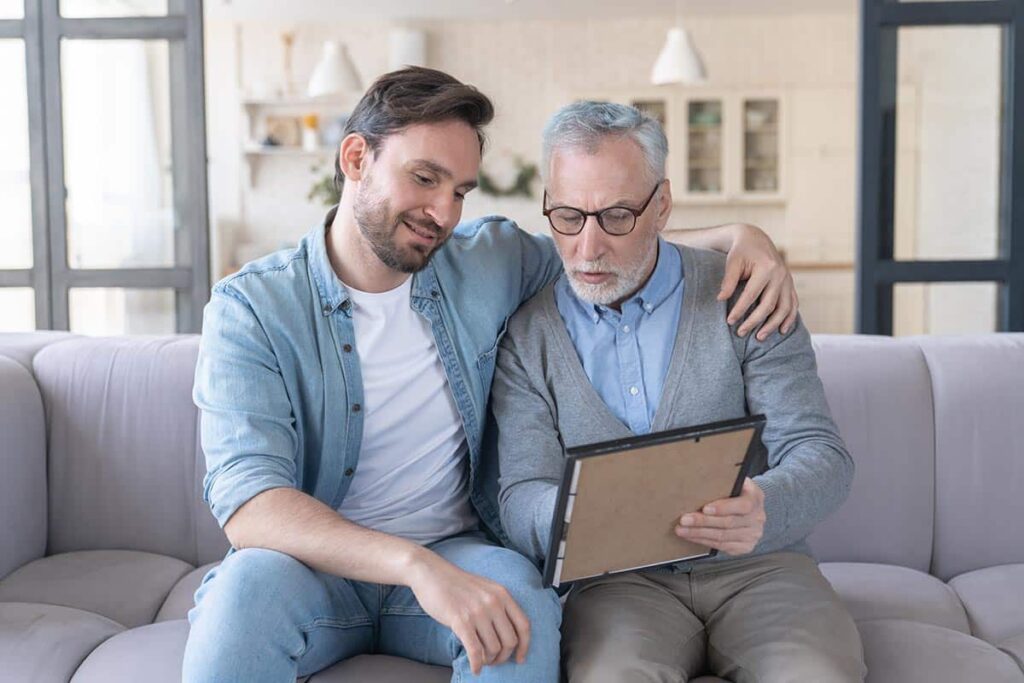 Pegasus Landing of Tanglewood | Caring loving adult son hugging embracing his old elderly senior father while he is showing his family photograph